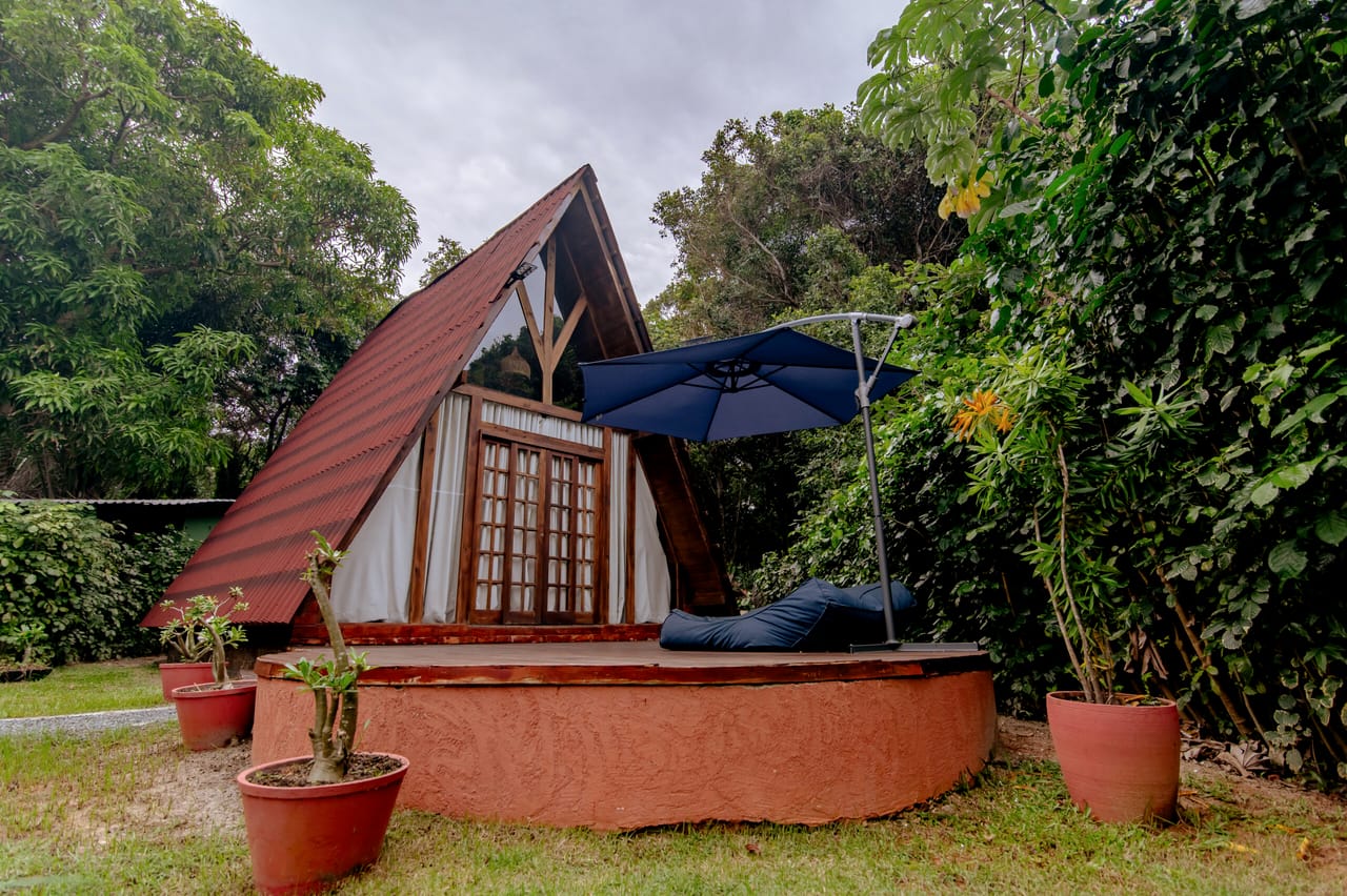 Cabana de madeira em formato de A entre árvores altas, com guarda-sol azul no deck